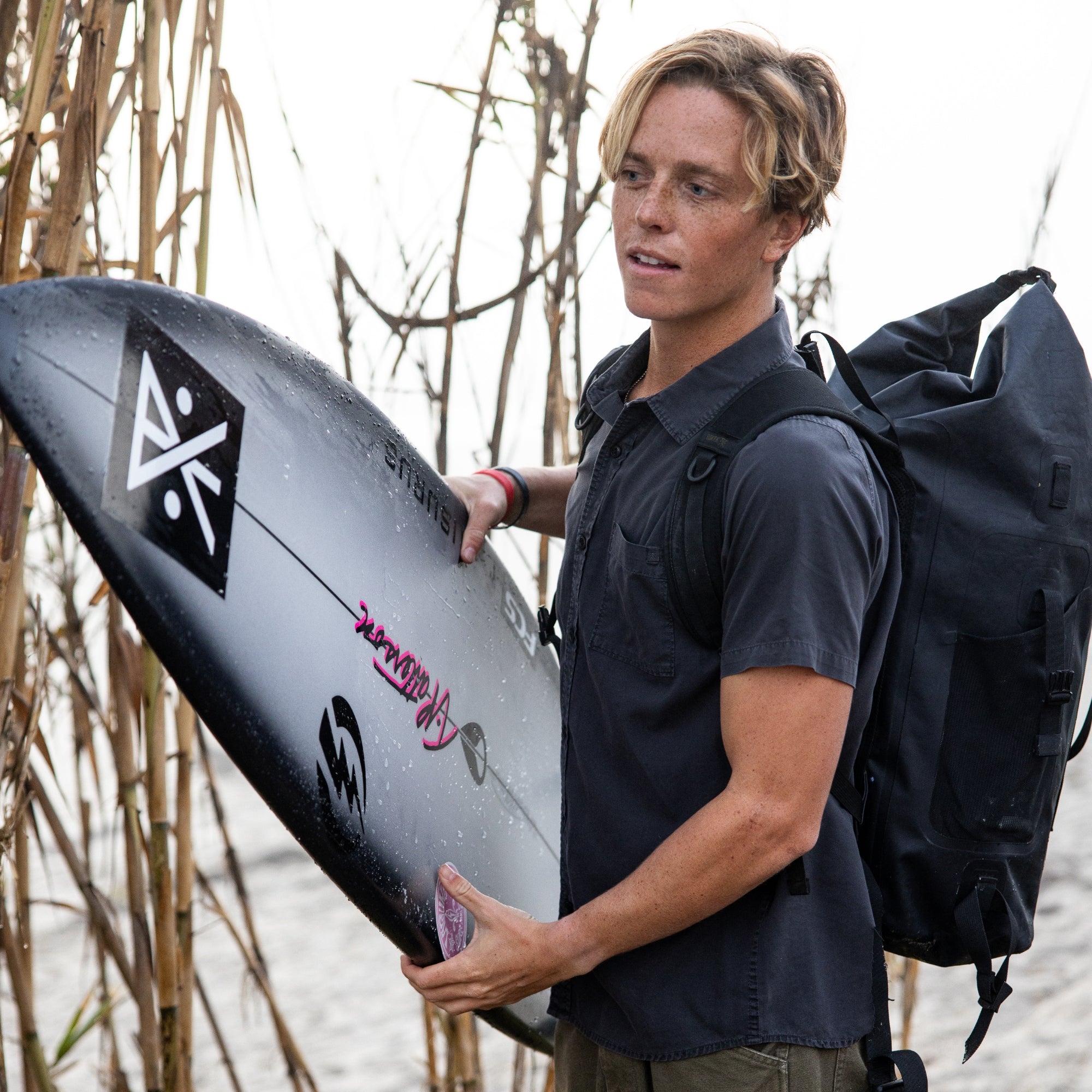 Man holding a surfboard with a Roark logo, standing outdoors near reeds.