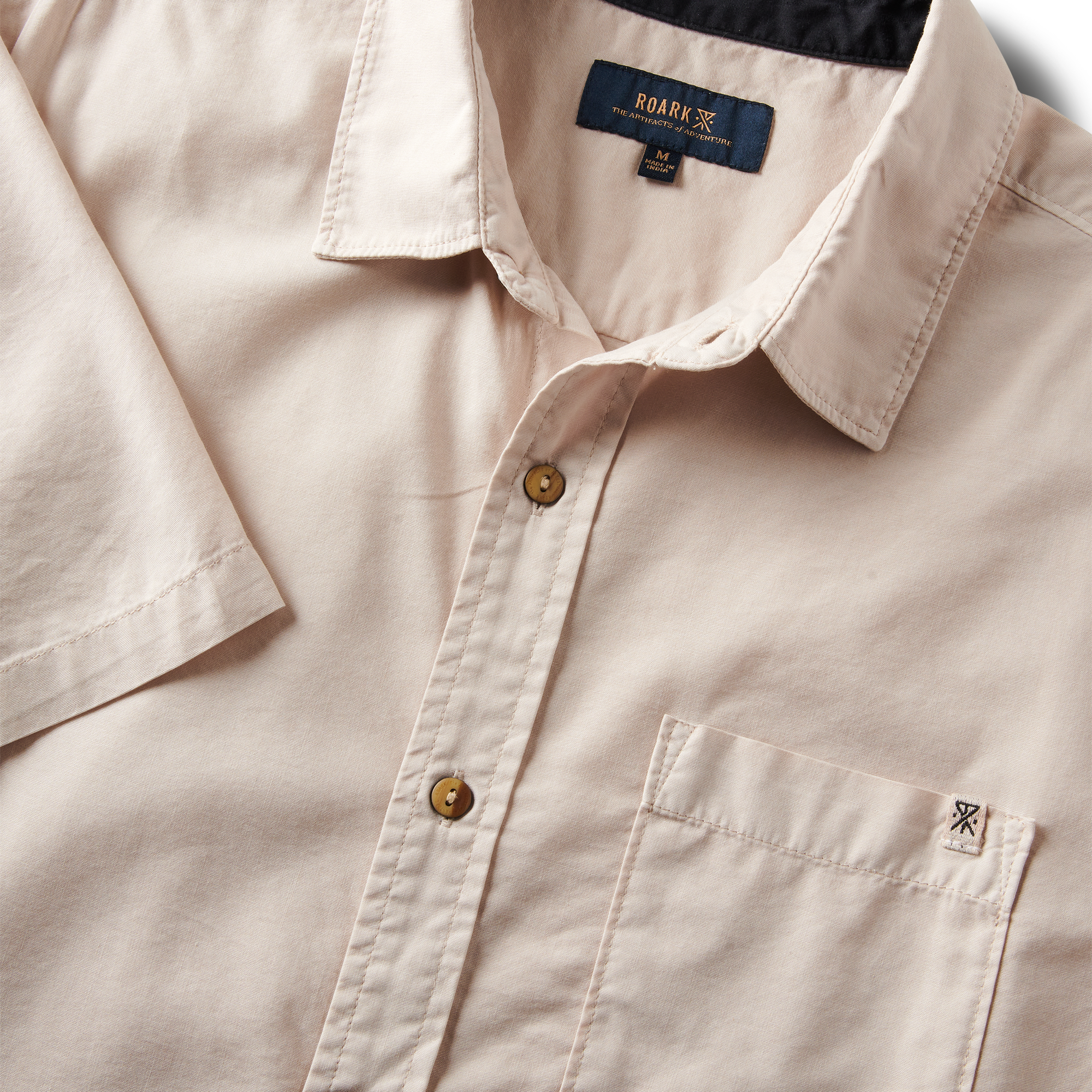 Close-up of the Roark Made To Fade Short Sleeve Button Up Shirt's collar, buttons, and chest pocket with embroidered logo in Washed Bone.