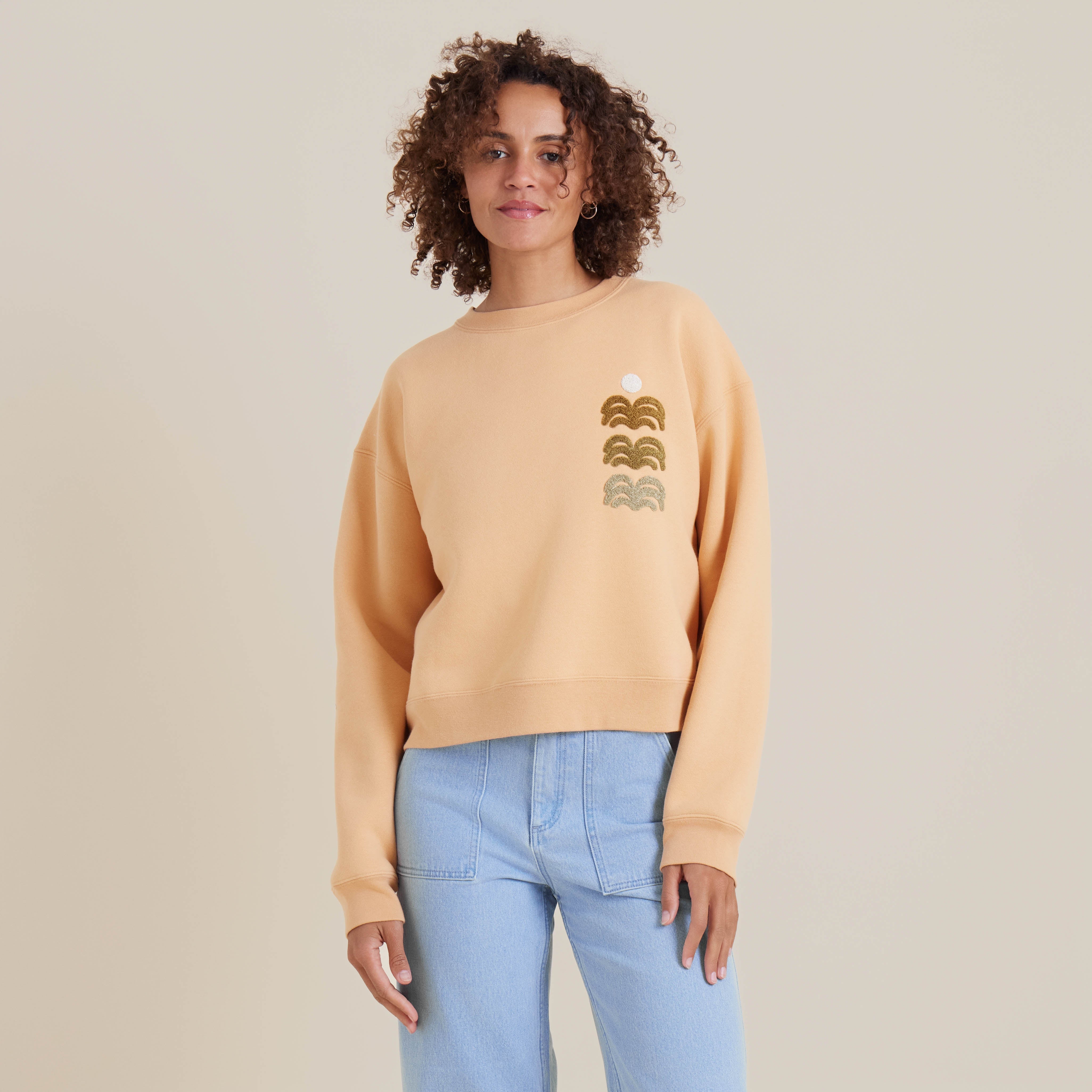 Model wearing a peach crewneck sweatshirt with a minimal wave graphic design, styled with light-wash jeans for a casual chic look.