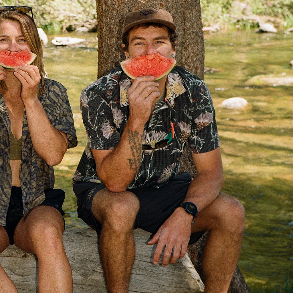 Two people sitting by a river, eating watermelon slices.