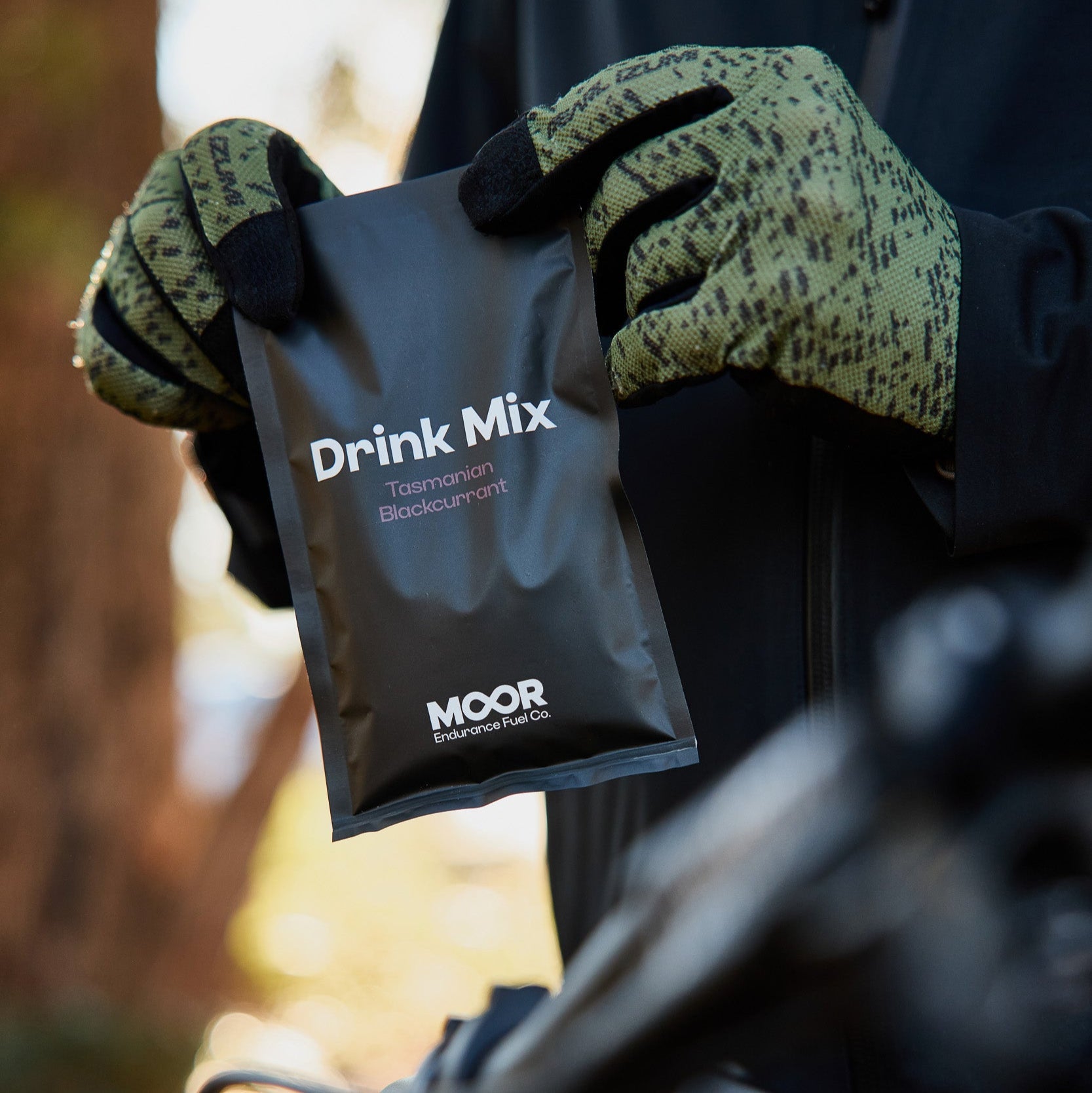 Person holding a 'Drink Mix' package with 'MOOR' branding, wearing gloves, against a blurred natural background.