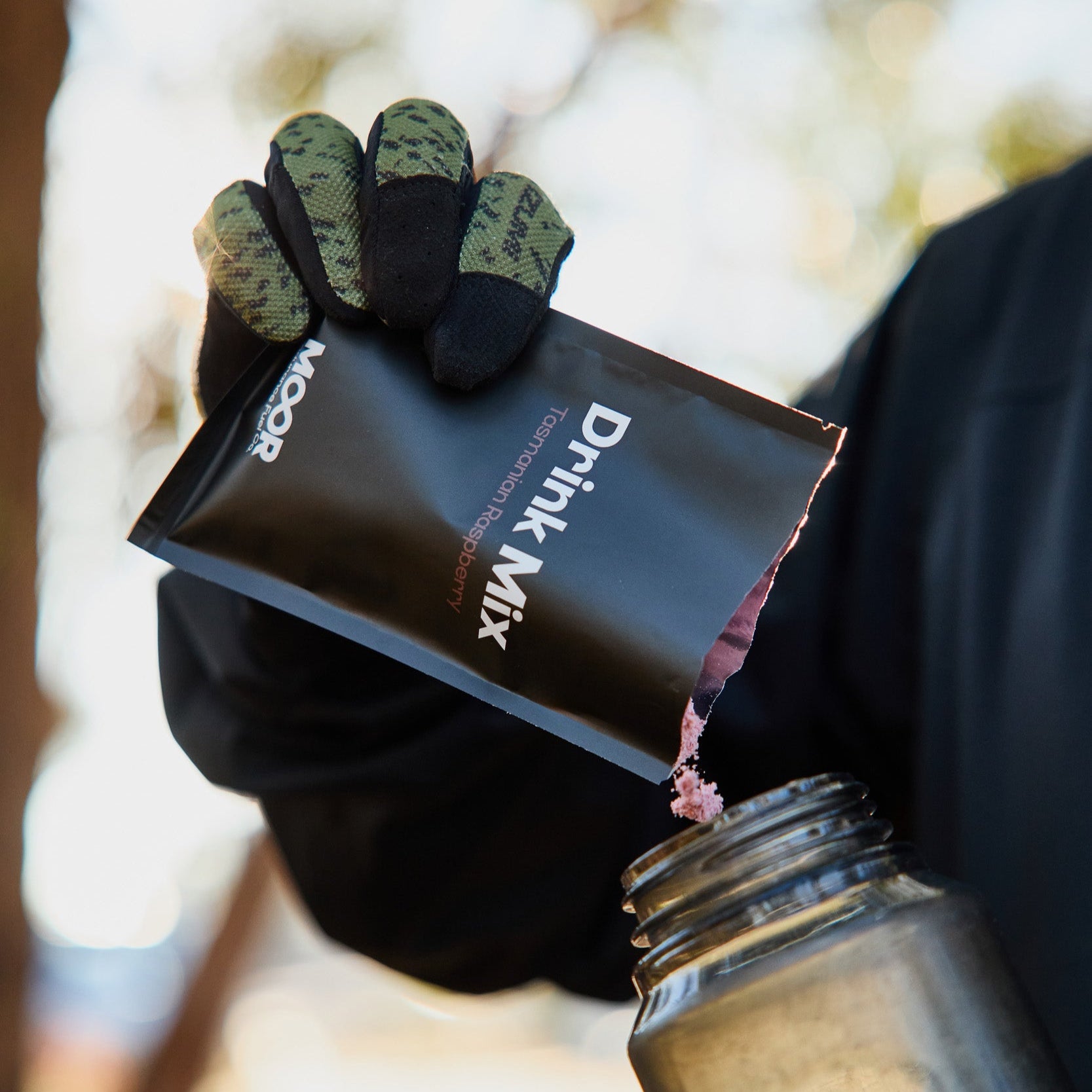 Person pouring a black container labeled 'Drink Mix' into a jar with a blurred outdoor background.