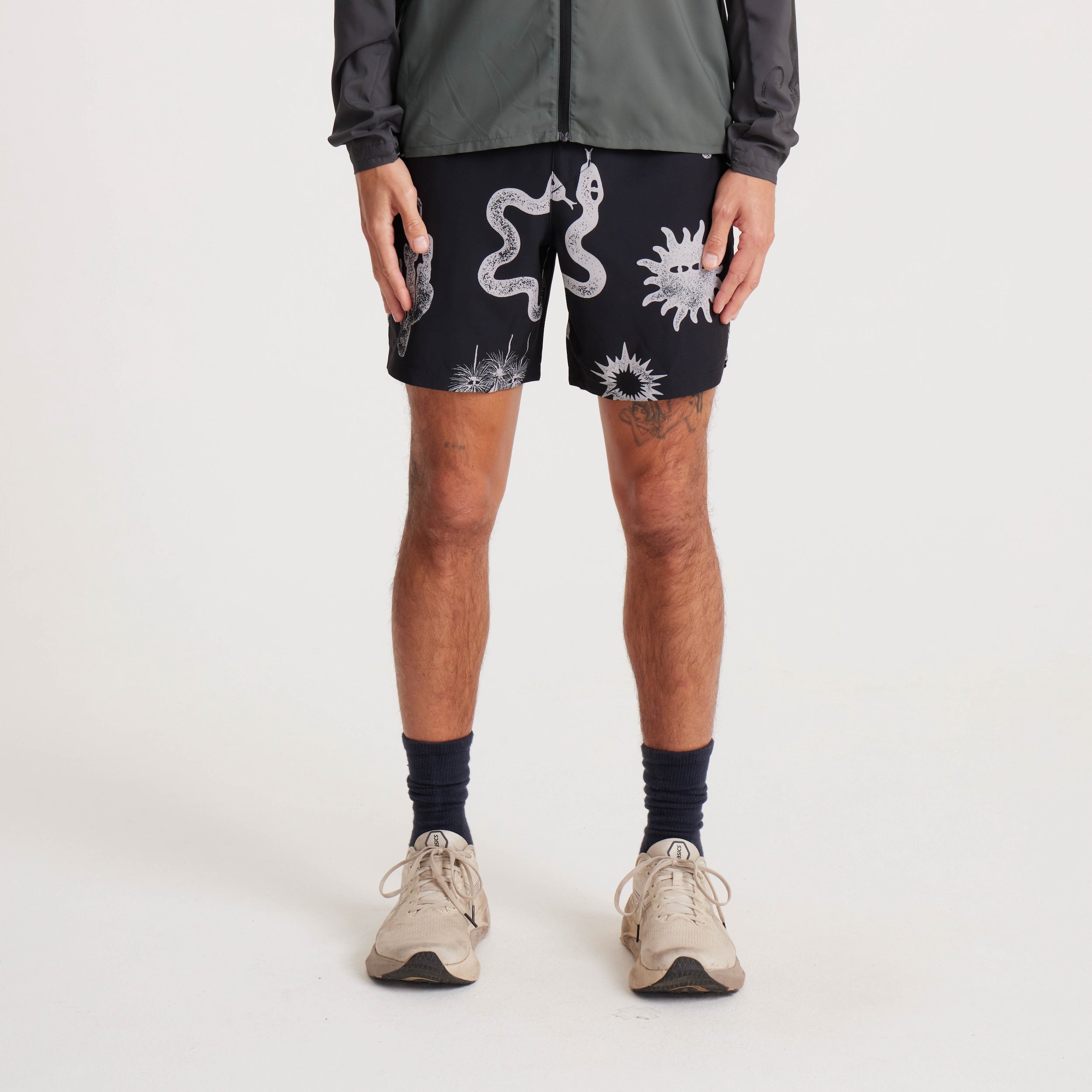 Model wearing black swim shorts with white graphic prints of snakes and desert-inspired patterns, styled with a lightweight jacket and sneakers for a casual summer look.
