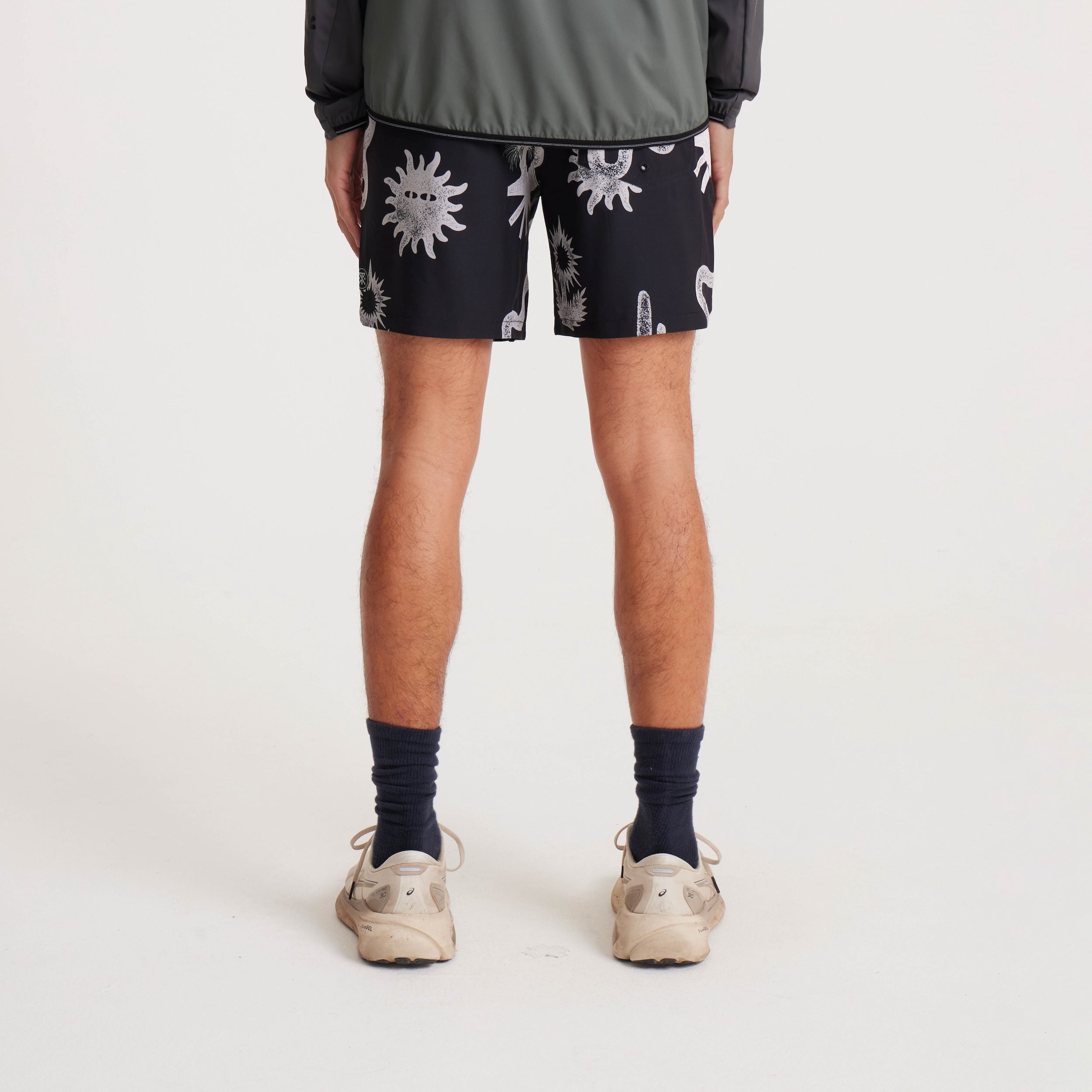 Back view of black swim shorts featuring white graphic desert-inspired designs, including snakes and abstract suns, paired with a casual jacket and sneakers.