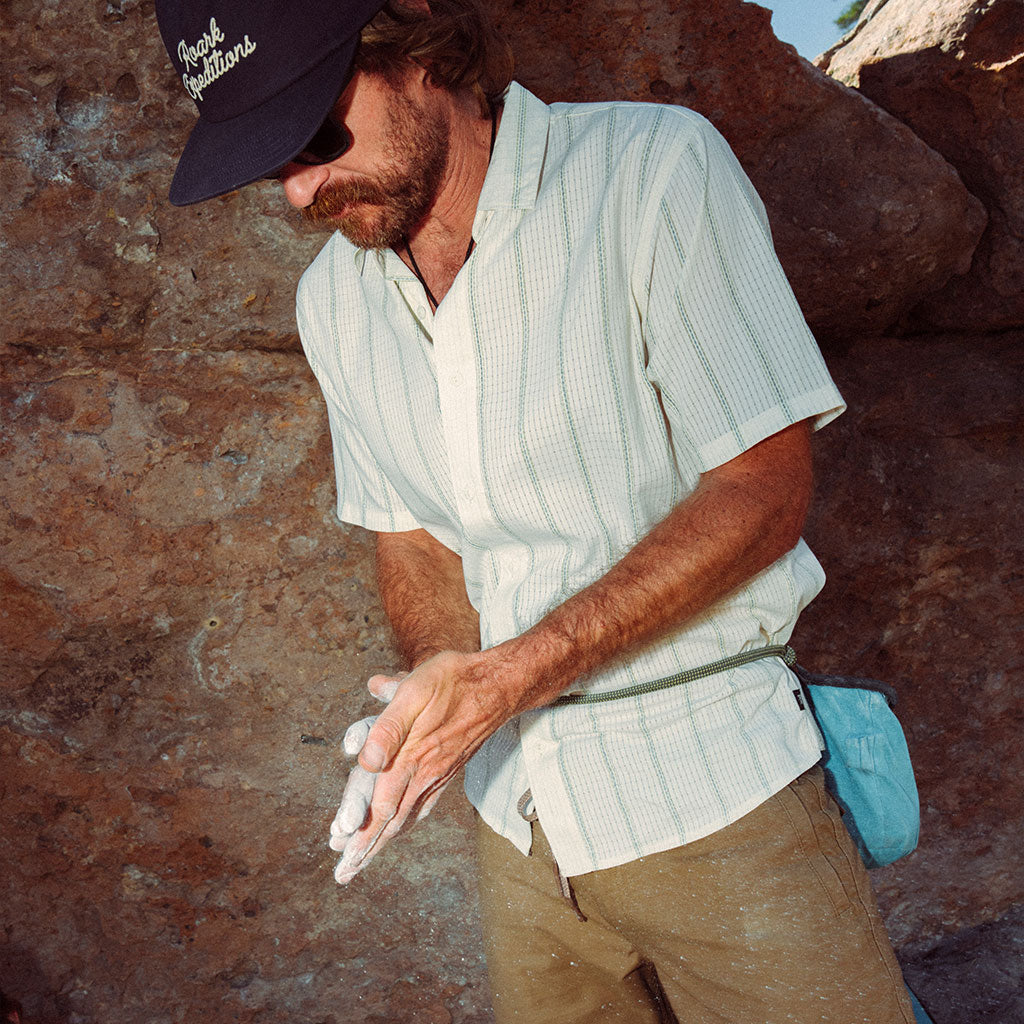 Man in outdoor setting wearing a cap and light shirt, standing against a rock wall.